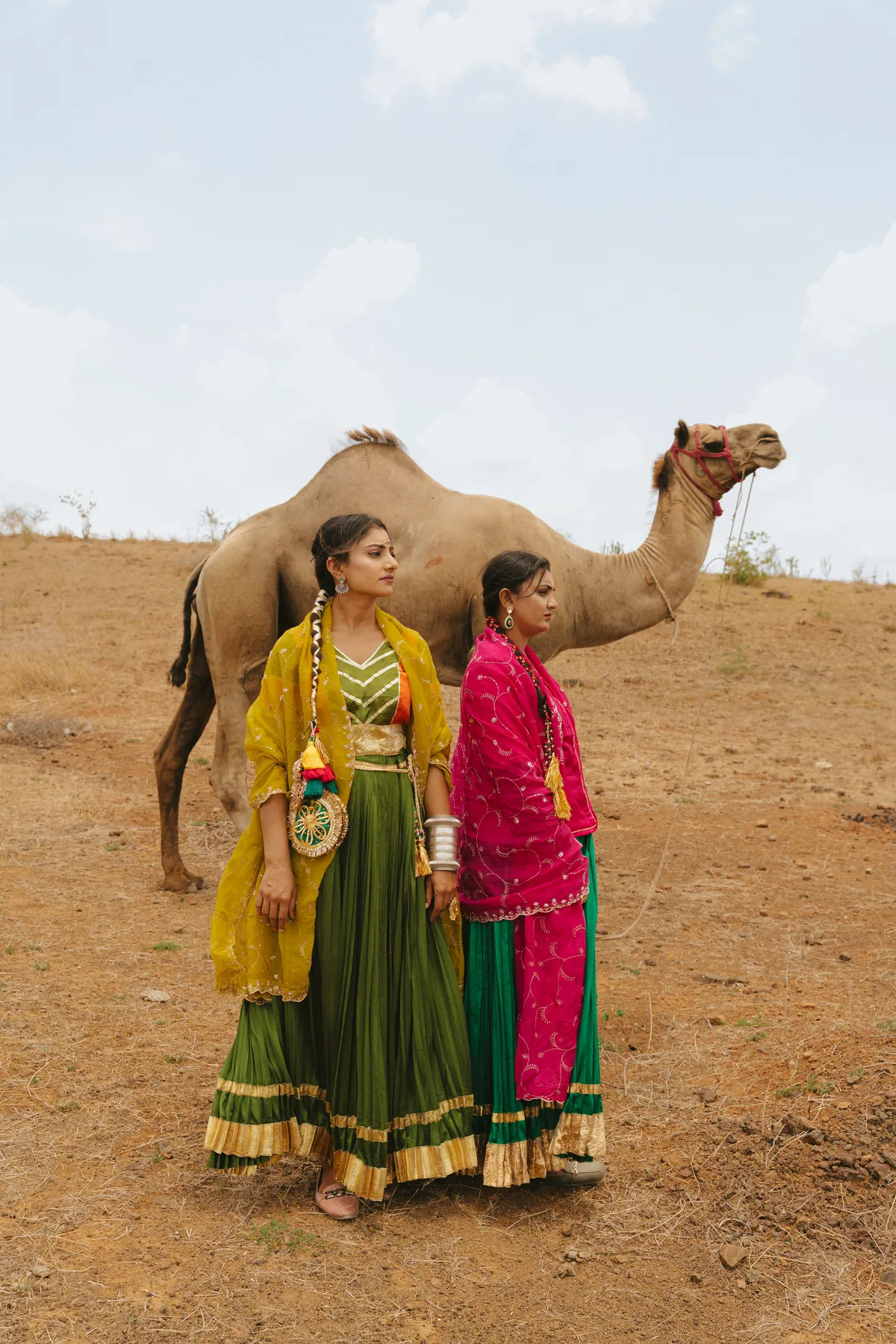 Two women in green skirts and colourful dupattas beside a camel